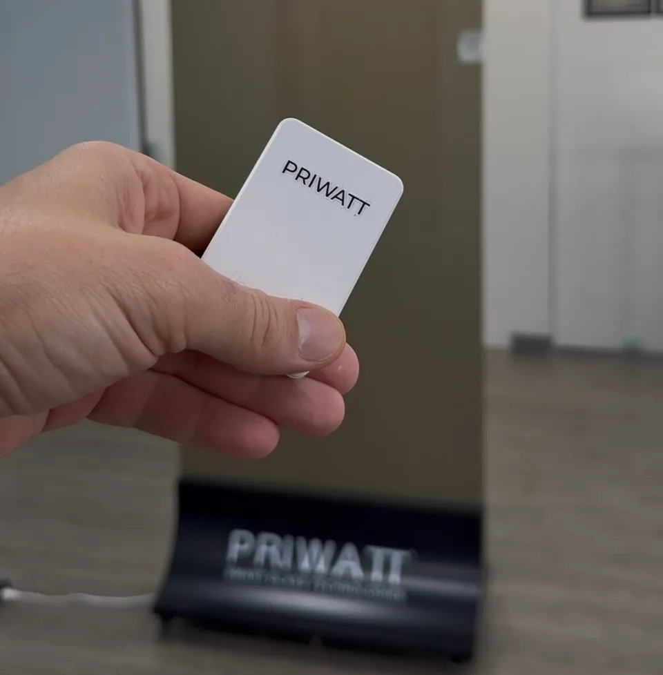 A hand holding a slim PriWatt remote in front of a switchable glass partition